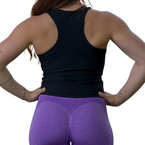 A person with long brown hair, wearing the LuxeForm Tank - Black and high-waisted purple leggings, stands hands on hips, back facing the camera against a white background.