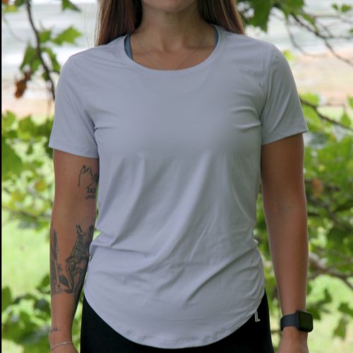 A woman with long brown hair, tattoos on her left arm, and a smartwatch wears the VeloraFlow Tee in Whisper Blue and black pants outdoors before blurred green trees.
