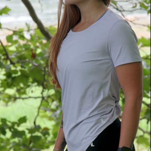 A woman with long brown hair, dressed in the VeloraFlow Tee - Whisper Blue, black leggings, and a smartwatch, stands outdoors before green trees and gazes to the side.