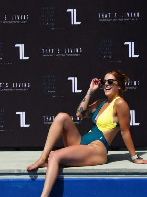 A woman in a Womens Vita One Swim Suit - Green/Yellow sits by the pool, smiling and adjusting her sunglasses, with a black backdrop behind her that displays the text “THAT’S LIVING.”.