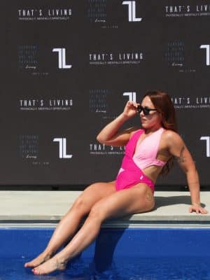 A woman in a Womens Vita One Swim Suit - Soft/Dark Pink sits at the pool's edge, dipping her feet in and adjusting her sunglasses. Behind her, a black backdrop displays "That's Living" and several logos.