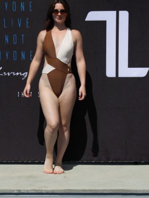 A woman stands barefoot by a pool in front of a black backdrop wearing the Womens Vita One Swim Suit in Mocka/White, paired with sunglasses and looking forward. White and blue text is displayed behind her.