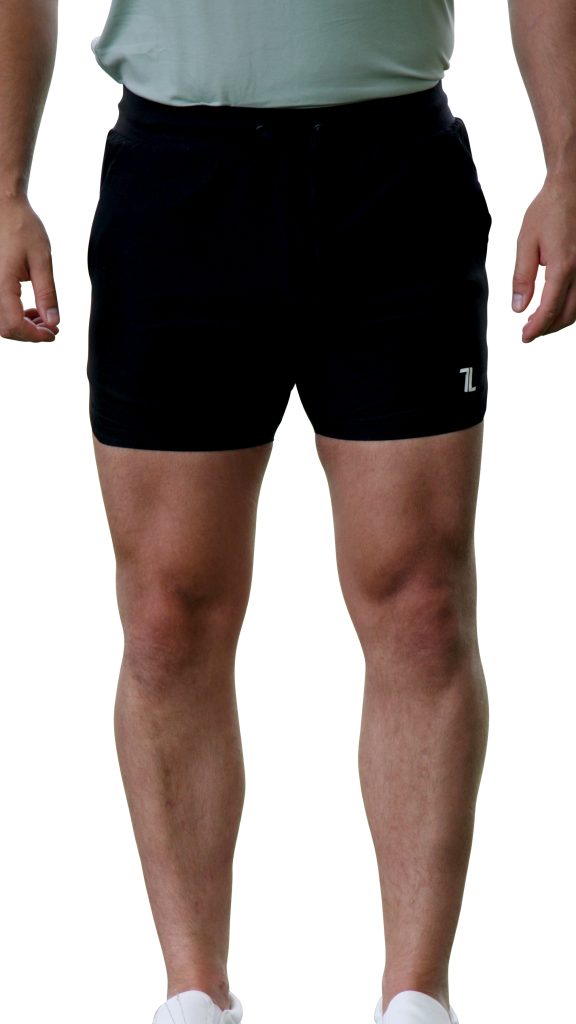 A person wearing a light green shirt, HYDRX Swim Shorts - Black, and white shoes stands with arms relaxed at their sides against a white background.