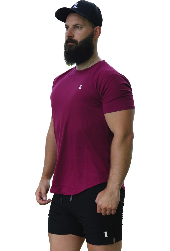 A bearded man in a black cap, Genesis Tee V2 - Maroon with tapered fit and scoop bottom, and black shorts stands against a white background, looking off to the side with his hands at his sides.