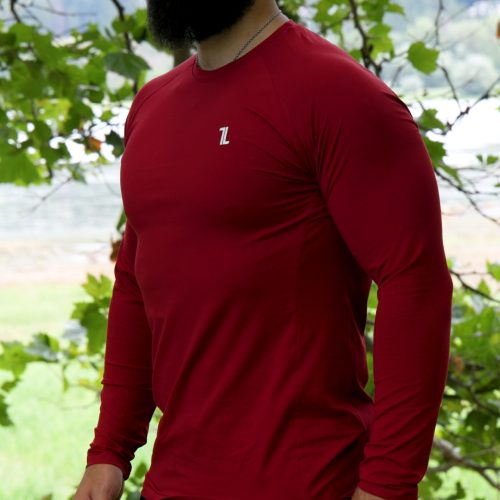 A bearded man wearing the FRMR Long sleeve - Dark Ember and a black cap stands outdoors among leafy trees, looking to the side.