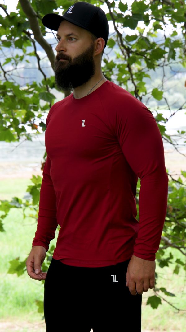 A bearded man wearing the FRMR Long sleeve - Dark Ember and a black cap stands outdoors among leafy trees, looking to the side.
