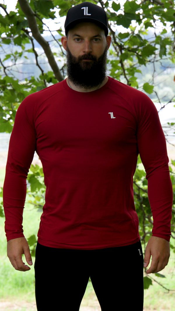 A bearded man in a red FRMR Long sleeve - Dark Ember and a black cap stands outdoors before leafy trees, looking directly at the camera.