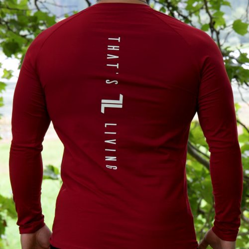 A man stands outdoors among trees, facing away in a red FRMR Long sleeve - Dark Ember men's performance shirt with "THAT'S LIVING" printed vertically on the back.