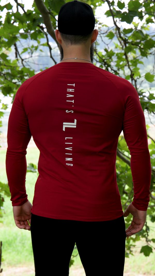 A man stands outdoors among trees, facing away in a red FRMR Long sleeve - Dark Ember men's performance shirt with "THAT'S LIVING" printed vertically on the back.