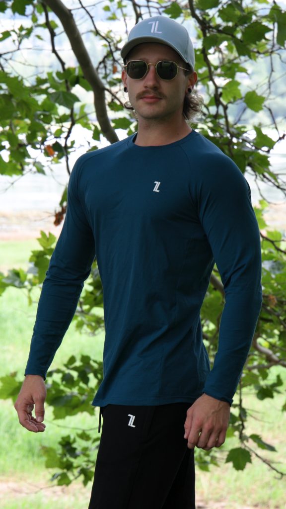 A man stands outdoors in front of leafy trees, looking at the camera with a neutral expression. He wears sunglasses, a gray cap, black pants, and the FRMR Long sleeve - Deep Ops gym shirt in teal.