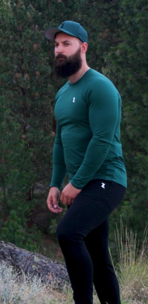 A bearded man in the FRMR Long Sleeve - Recon Pine, black pants, and a green cap stands outdoors among pine trees and grass, gazing to the side.