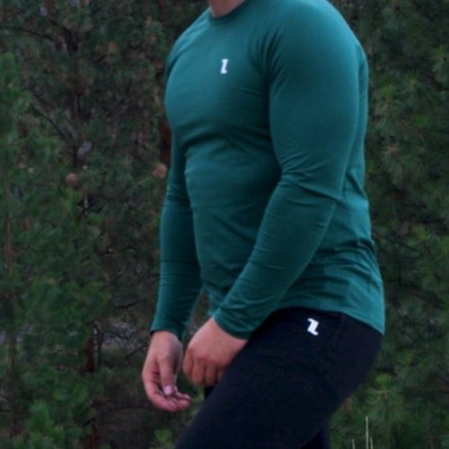 A bearded man in the FRMR Long Sleeve - Recon Pine, black pants, and a green cap stands outdoors among pine trees and grass, gazing to the side.