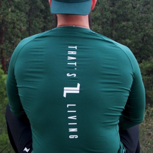 A person wearing the FRMR Long Sleeve - Recon Pine and a matching hat sits outdoors with their back to the camera. "THAT'S L LIVING" is printed in white on the shirt, set against a forest and rocky hillside backdrop.