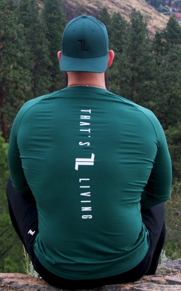 A person wearing the FRMR Long Sleeve - Recon Pine and a matching hat sits outdoors with their back to the camera. "THAT'S L LIVING" is printed in white on the shirt, set against a forest and rocky hillside backdrop.