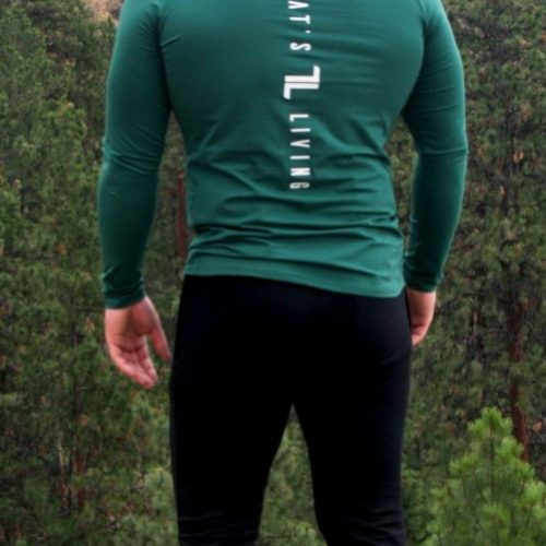 A man wearing the FRMR Long Sleeve - Recon Pine stands outdoors with his back to the camera, overlooking a forested area and rocky hillside. He pairs the shirt with black pants, white shoes, and a green cap.