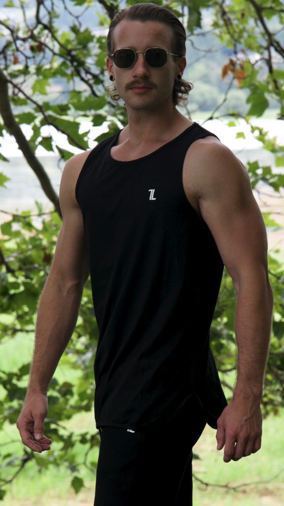 A man in sunglasses wears the Genesis Tank V2 - Black with the TL logo, standing outdoors among green trees and looking confidently at the camera.