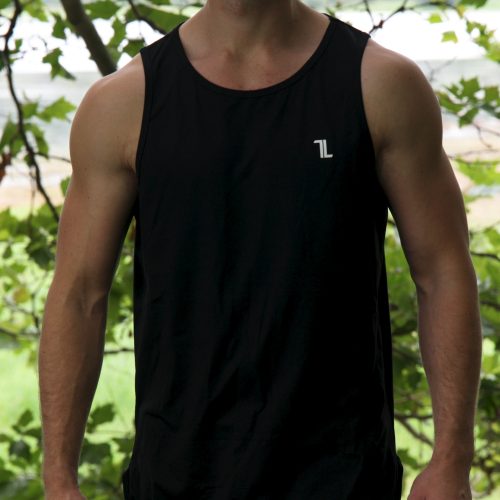 A man with sunglasses, a mustache, and long hair stands outdoors among leafy trees, wearing the black Genesis Tank V2 and black pants, looking straight at the camera.