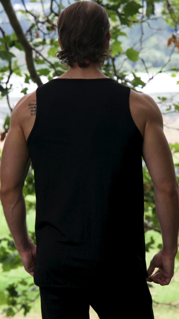 A person with medium-length hair stands outdoors facing away, wearing the Genesis Tank V2 - Black. The minimalist men’s performance tank features a TL logo and reveals part of a tattoo on their upper left shoulder amid greenery.
