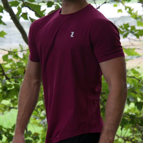 A man with long hair and a mustache stands outdoors before leafy trees, wearing sunglasses, black pants, and the Genesis Tee V2 in maroon—a men’s performance t-shirt with a tapered fit and scoop bottom.