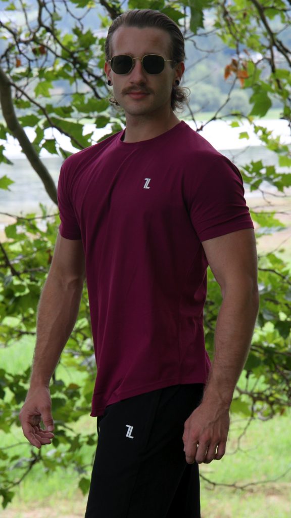A man with long hair and a mustache stands outdoors before leafy trees, wearing sunglasses, black pants, and the Genesis Tee V2 in maroon—a men’s performance t-shirt with a tapered fit and scoop bottom.