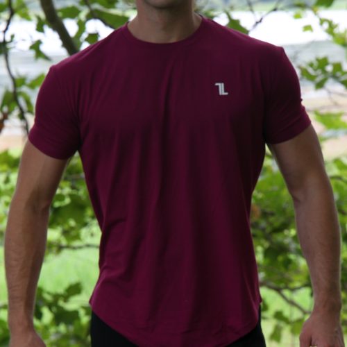 A person stands outdoors in front of greenery, smiling slightly with arms relaxed at their sides, wearing sunglasses, black pants, and the Genesis Tee V2 - Maroon men's performance t-shirt with a tapered fit and scoop bottom.