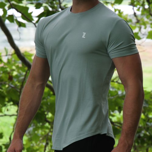 A man with a mustache and sunglasses stands outside by leafy trees, wearing the Genesis Tee V2 in Cool Mint—a light green men’s performance t-shirt with tapered fit, scoop bottom, and a small "7L" logo—paired with black pants.