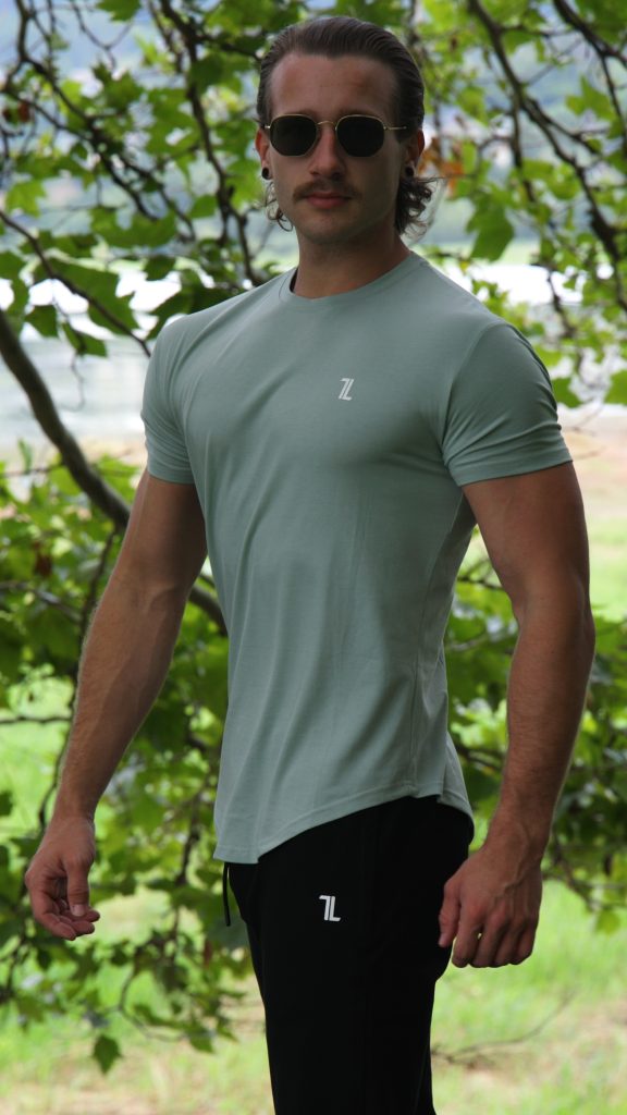 A man with a mustache and sunglasses stands outside by leafy trees, wearing the Genesis Tee V2 in Cool Mint—a light green men’s performance t-shirt with tapered fit, scoop bottom, and a small "7L" logo—paired with black pants.