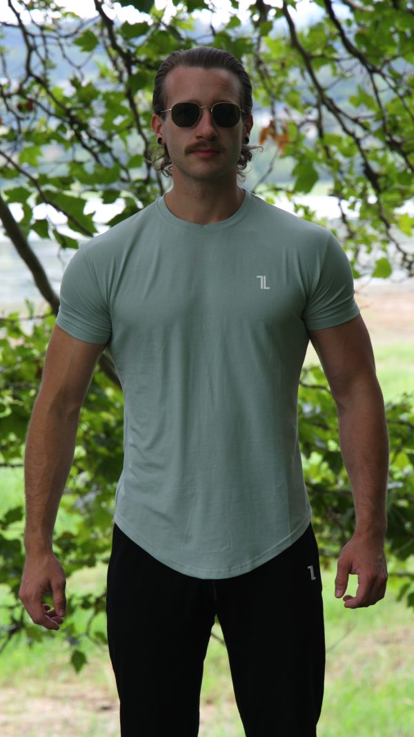 A man with long hair and sunglasses wears the Genesis Tee V2 in Cool Mint—a men’s performance shirt with a tapered fit and scoop bottom—and black pants, standing outdoors before leafy green trees, looking at the camera.