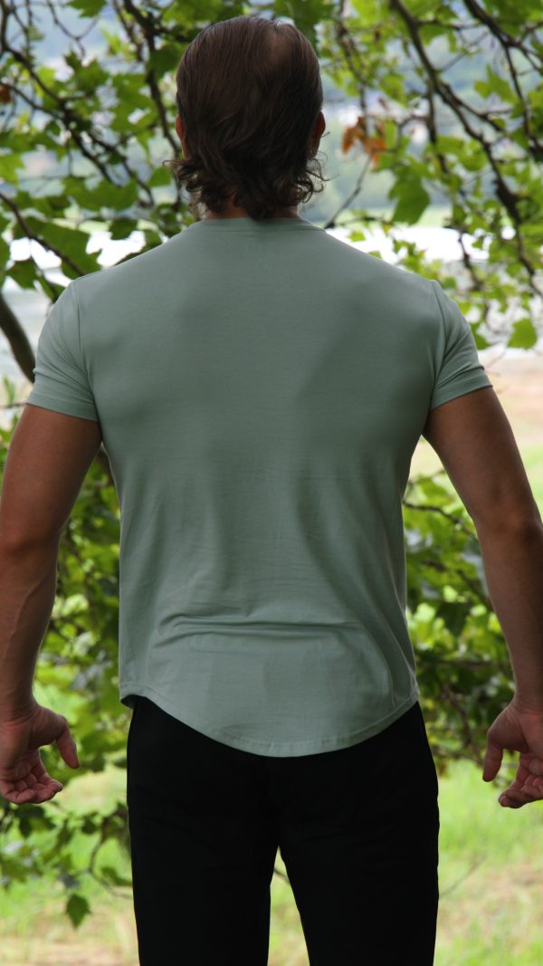 A person with medium-length brown hair, wearing the Genesis Tee V2 - Cool Mint (men’s performance t-shirt with tapered fit and scoop bottom) and black pants, stands outdoors with their back to the camera amid lush green foliage.