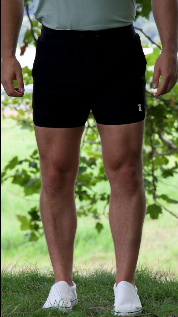 A person standing outdoors on grass with green foliage in the background, showing only their lower torso and legs, is wearing a light-colored shirt, white shoes, and HYDRX Swim Shorts - Black.