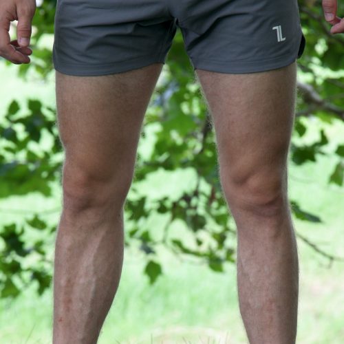A person stands outdoors on grass, wearing HYDRX Swim Shorts - Tidal Ash, a blue long-sleeve shirt, and black shoes. Green leaves and blurred grass form the background.