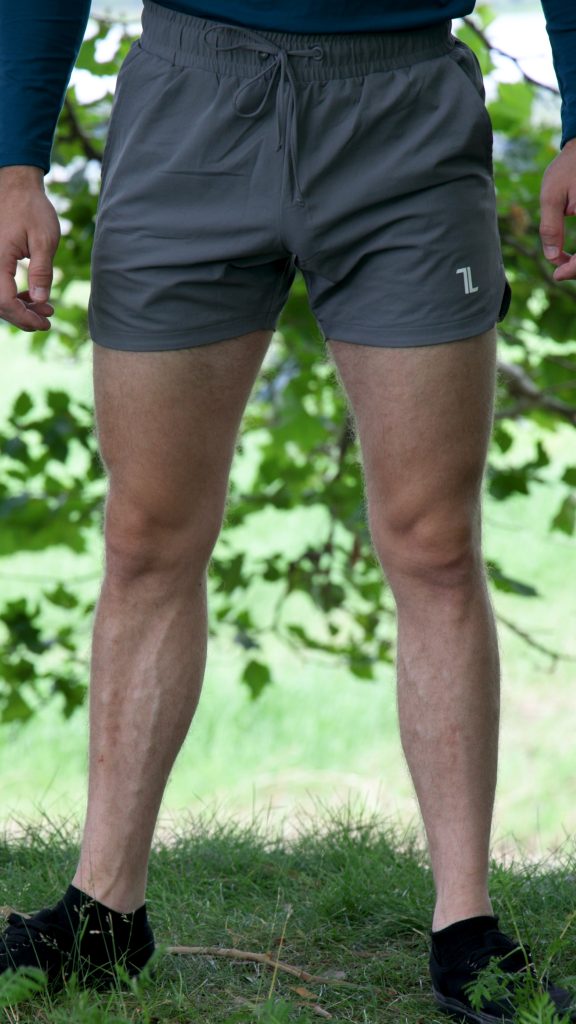 A person stands outdoors on grass, wearing HYDRX Swim Shorts - Tidal Ash, a blue long-sleeve shirt, and black shoes. Green leaves and blurred grass form the background.