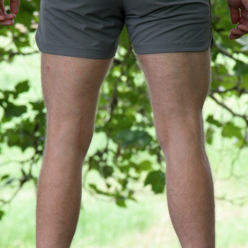 A person stands outdoors on grass, seen from behind, wearing HYDRX Swim Shorts - Tidal Ash with a dark top and black shoes, surrounded by leafy green trees.