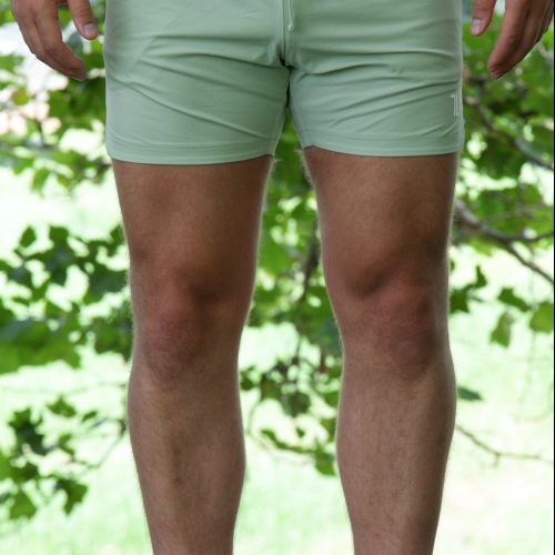 A person wearing HYDRX Swim Shorts - Waveleaf in sage green and white slip-on shoes stands on grass outdoors, with leafy green trees in the background.