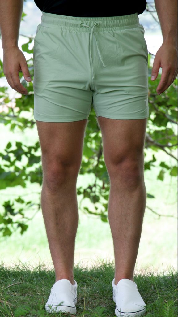 A person wearing HYDRX Swim Shorts - Waveleaf in sage green and white slip-on shoes stands on grass outdoors, with leafy green trees in the background.