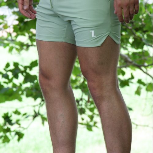 A person stands outdoors on grass in HYDRX Swim Shorts - Waveleaf (light green with white logo) and white slip-on shoes, surrounded by green foliage and sunlight. Their face is not visible.