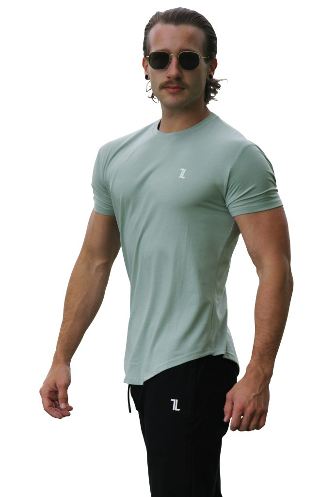 A man wearing sunglasses, black pants, and a light green Genesis Tee V2 - Cool Mint with tapered fit and scoop bottom stands facing forward with one arm bent. Both the tee and pants have a small white "7L" logo. The background is white.