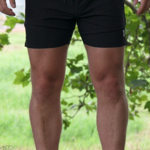 Outdoors on grass, a person wears Rush Shorts - Black and white shoes. Green leaves and blurred scenery form the background, with only the legs and lower torso visible.