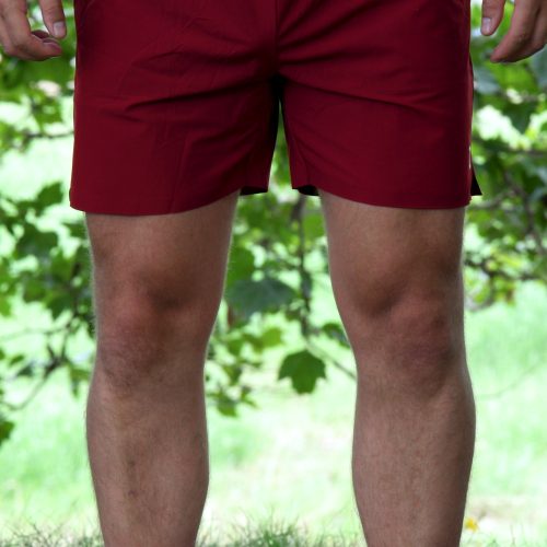 A person stands outdoors wearing Rush Shorts - Crimson Force with white shoes. Their hands rest by their sides, surrounded by green grass and leafy trees in the background.