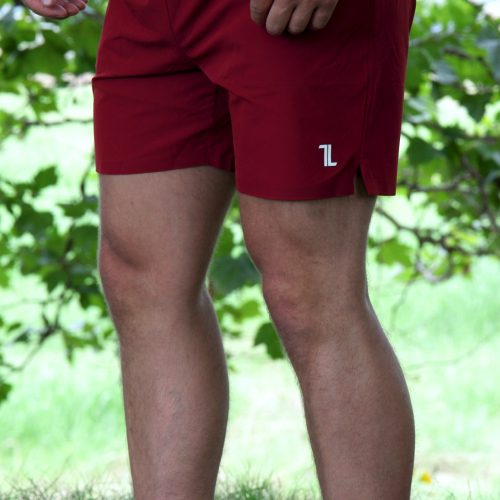 A person stands on grass wearing Rush Shorts - Crimson Force, featuring a small white logo and paired with white shoes. Their upper body is mostly out of frame, with green leaves in the background.