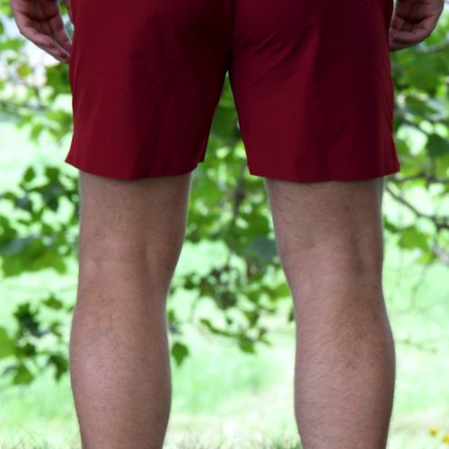 Outdoors on grass, a person faces away wearing Rush Shorts - Crimson Force and white shoes, with green leaves and branches blurred in the background.