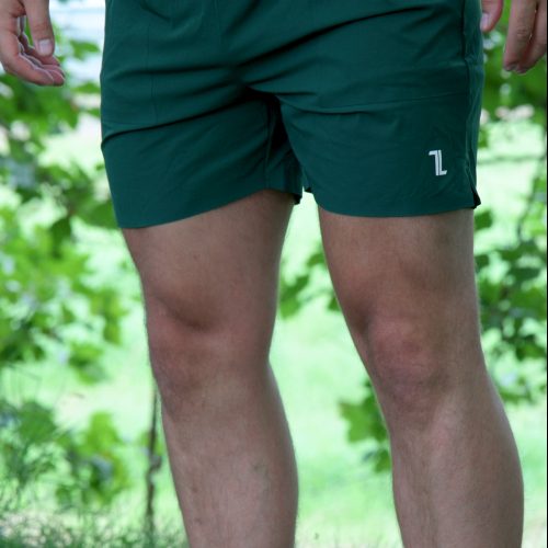 A person stands on grass wearing Rush Shorts - Urban Forest and white shoes, with trees and greenery blurred in the background and most of their upper body out of frame.