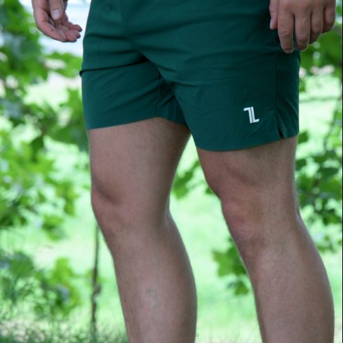 A person stands on grass wearing the Rush Shorts - Urban Forest, featuring a white logo on the left leg, paired with a black shirt and white slip-on shoes. Green-leaved trees are visible in the background.