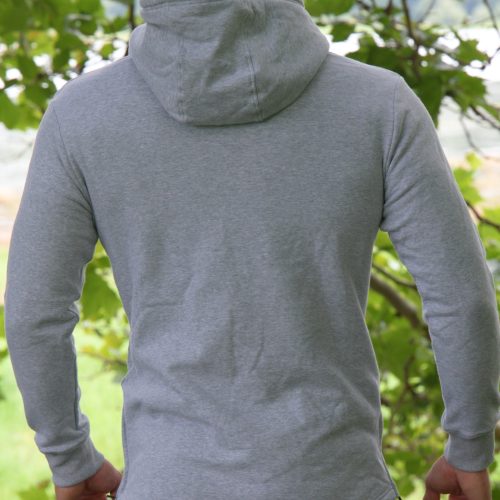 A person with long hair stands outdoors, back to the camera, surrounded by green foliage and trees, wearing the SPEKTR Hoodie - InfraGrey.