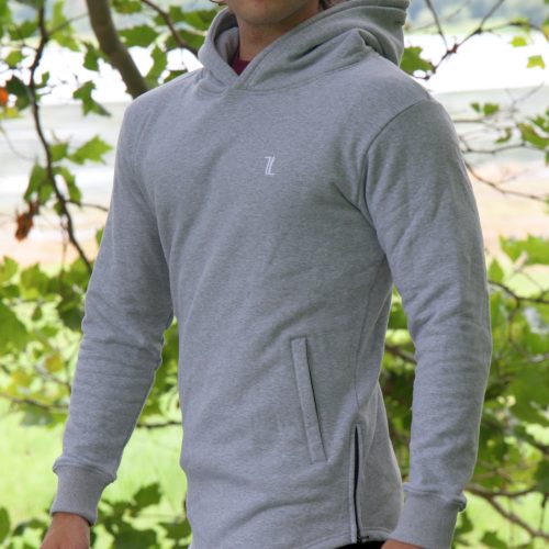A man stands outdoors before green trees, looking at the camera while wearing sunglasses, black pants, and the SPEKTR Hoodie in InfraGrey.