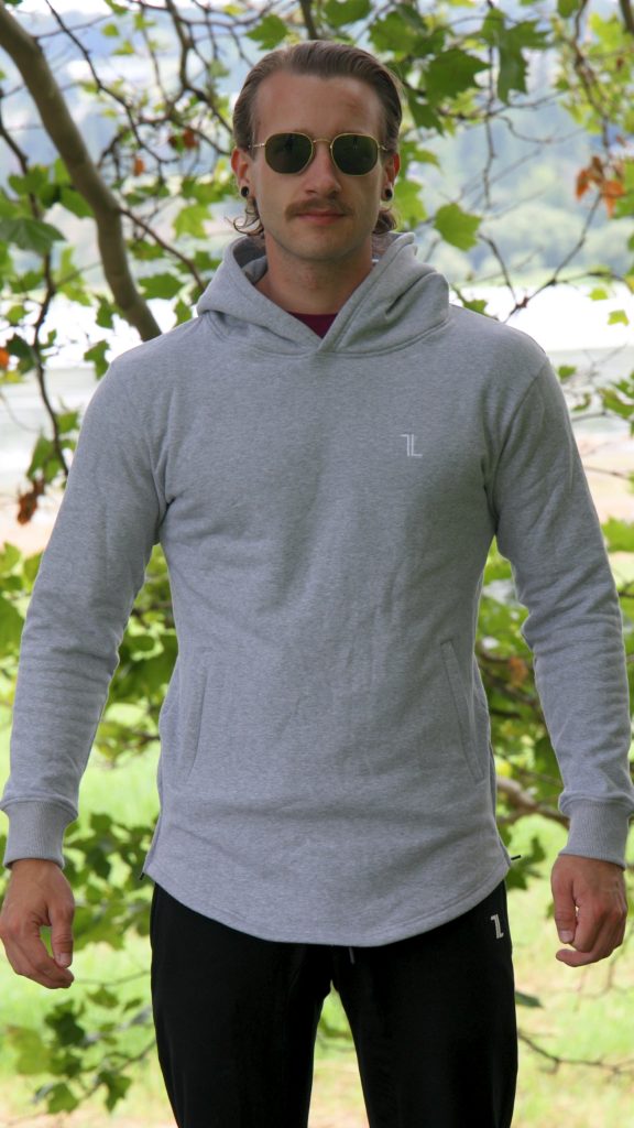 A man wearing the SPEKTR Hoodie - InfraGrey and sunglasses stands outdoors in front of leafy green trees, with blurred grass and sky in the background.