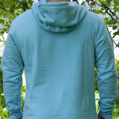 Wearing the SPEKTR Hoodie in Sky Blue and a matching cap, a person stands outdoors with their back to the camera, surrounded by green leaves and trees.