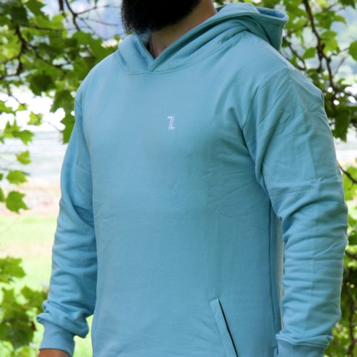 A bearded man stands outdoors before leafy green trees, wearing the SPEKTR Hoodie in Sky Blue and a matching cap, looking off to the side.