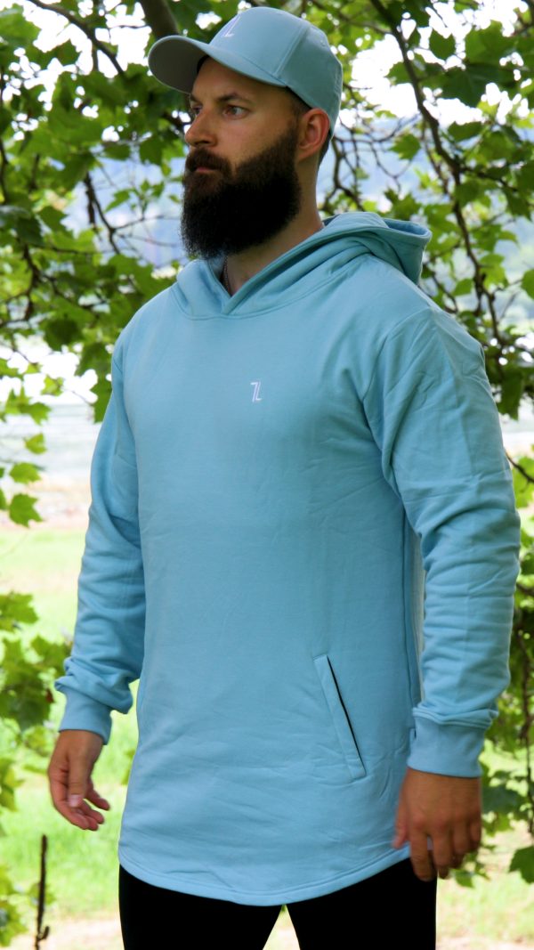 A bearded man stands outdoors before leafy green trees, wearing the SPEKTR Hoodie in Sky Blue and a matching cap, looking off to the side.