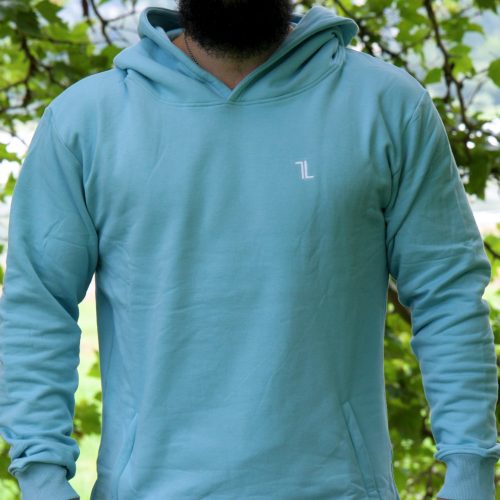 A bearded man stands outdoors before leafy trees, wearing a SPEKTR Hoodie in sky blue for high performance and a matching cap, both featuring a small white logo on the chest and hat.